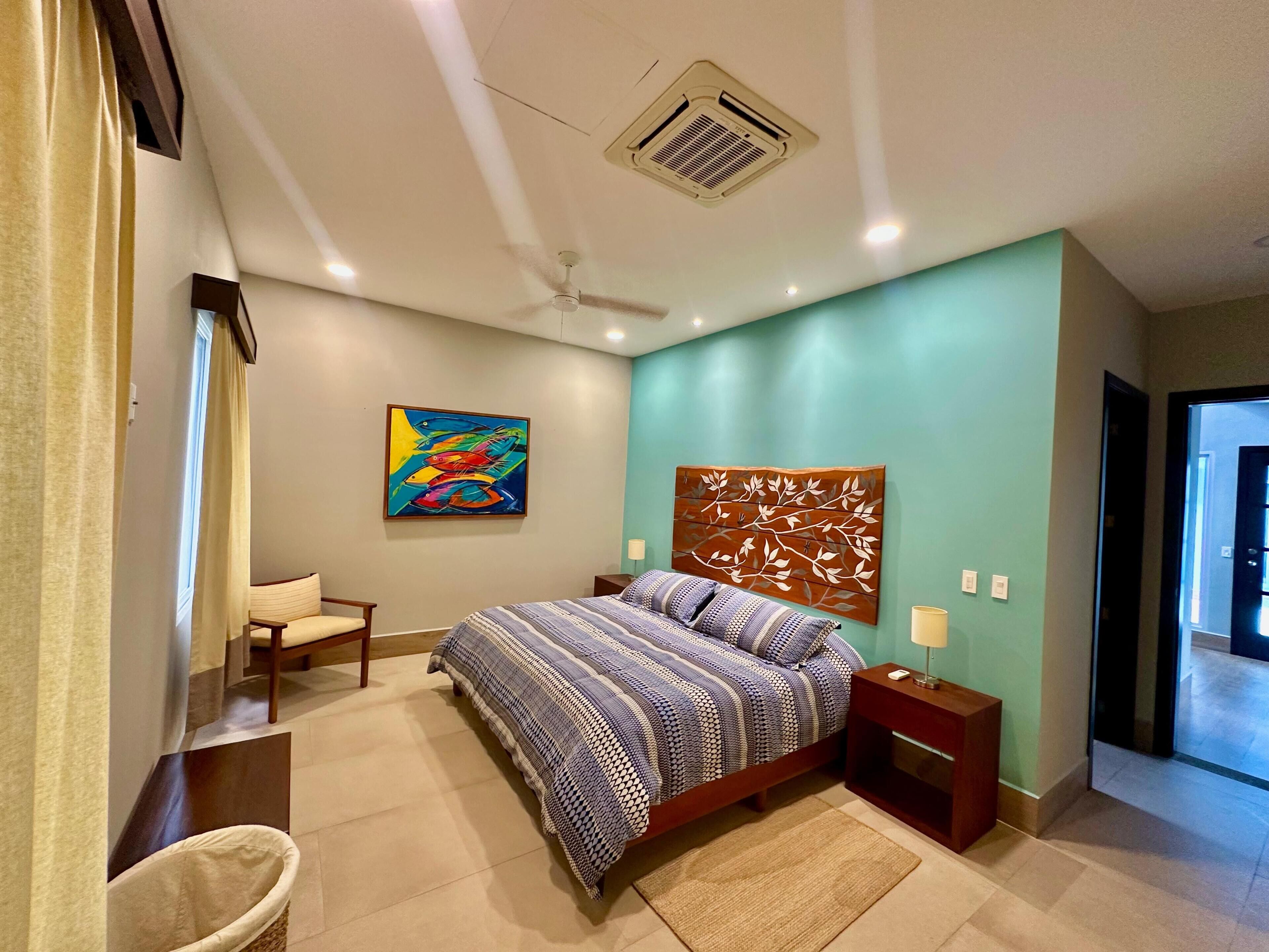 Un dormitorio con cama, mesita de noche, una silla, ventilador de techo y una obra de arte en la pared.