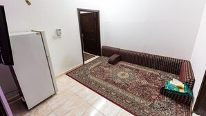 Une petite pièce avec un tapis à motifs, un canapé, un réfrigérateur et une porte qui donne sur une autre pièce.