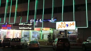 A building with vertical green lighting, multiple shops with signage, and parked cars in front.