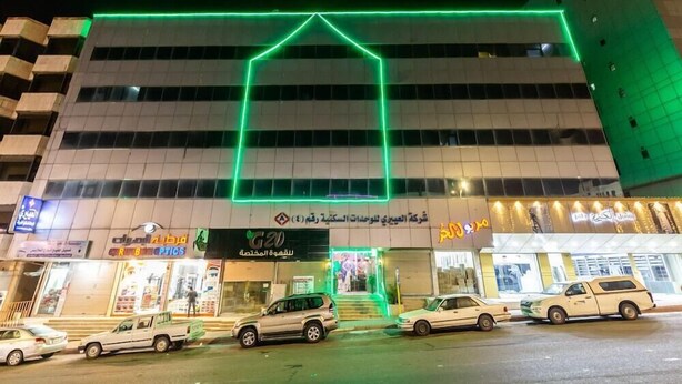 Un edificio con un contorno de neón verde que dibuja una casa, varias tiendas y coches estacionados al frente.