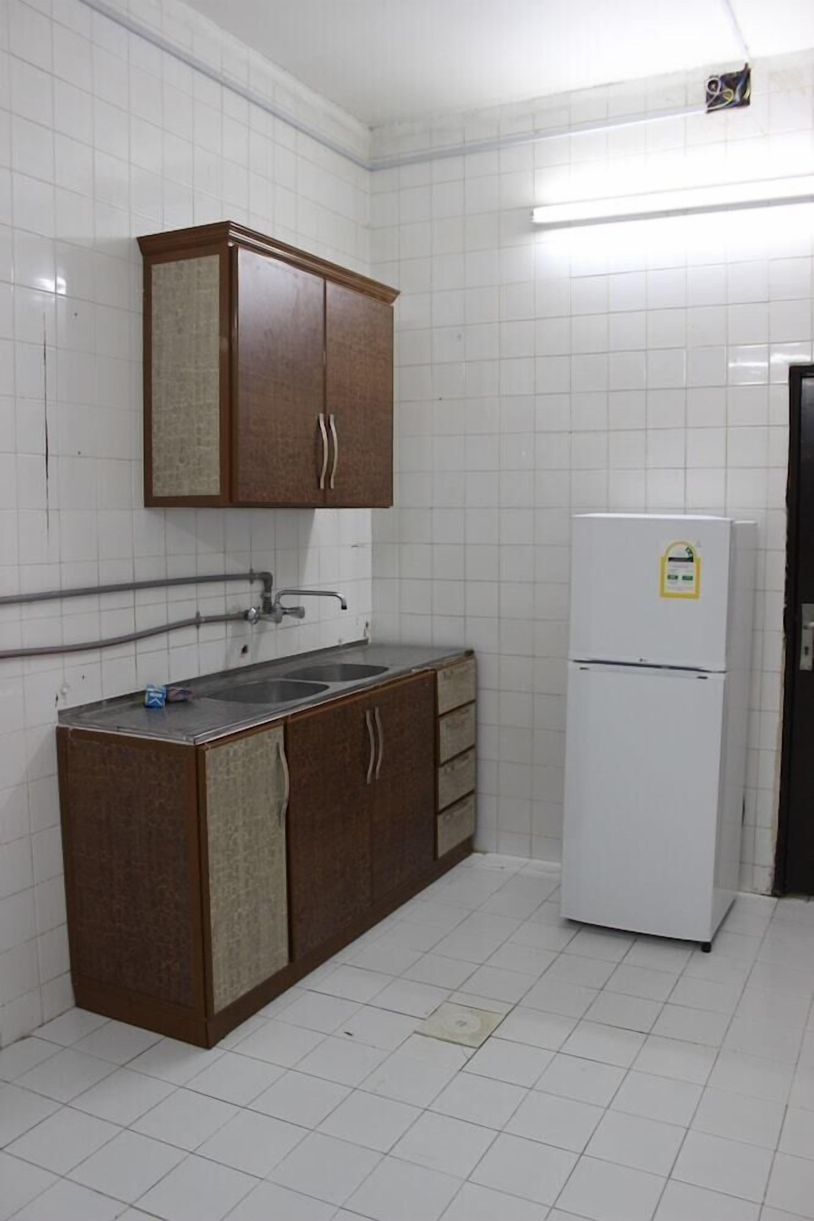 Une petite cuisine aux murs recouverts de carreaux blancs, un meuble en bois à deux portes, un évier et un réfrigérateur blanc.