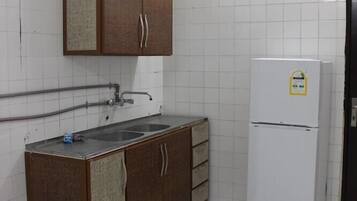 Une petite cuisine aux murs recouverts de carreaux blancs, un meuble en bois à deux portes, un évier et un réfrigérateur blanc.