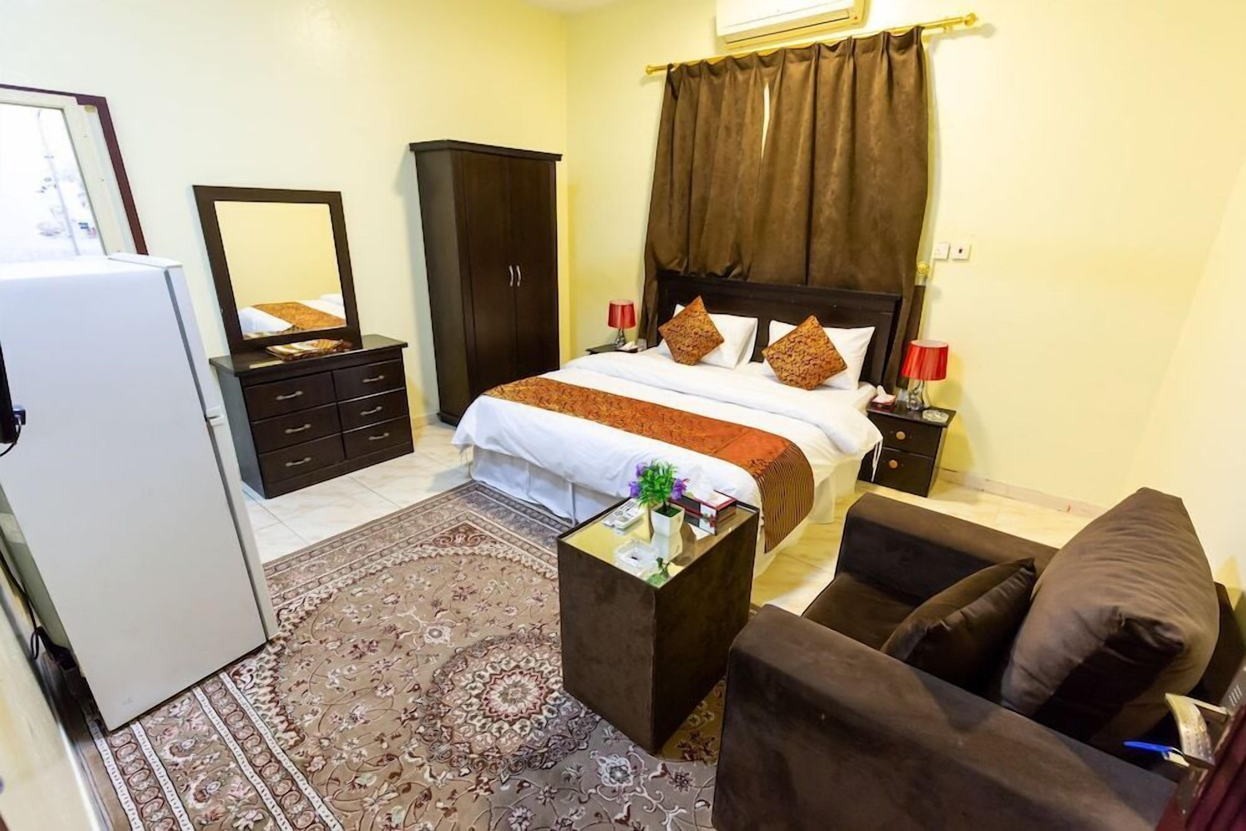 Une chambre d’hôtel comprenant un lit, une armoire, un bureau avec une lampe, un canapé et un tapis à motifs.