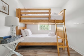 Uma cama beliche com escada, uma mesinha de cabeceira com luminária e uma janela com vista para o verde.