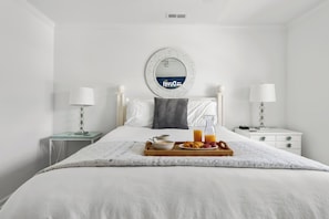 A hotel room with a neatly made bed, a breakfast tray with fruits and juice, a bedside table with a lamp, and a wall-mounted mirror. - Coastal Townhouse | Minutes from Everything (Mt Pleasant)