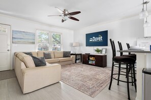 Un salon avec un canapé beige, une table basse, un tapis, un ventilateur de plafond, un téléviseur et un coin bar avec des tabourets.