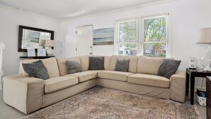 Ein beiges Ecksofa mit mehreren Kissen, ein gemustertes Teppich, eine weiße Tür, ein Gemälde und ein Fenster mit Jalousien.