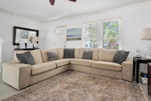 Ein beiges Ecksofa mit mehreren Kissen, ein gemustertes Teppich, eine weiße Tür, ein Gemälde und ein Fenster mit Jalousien.