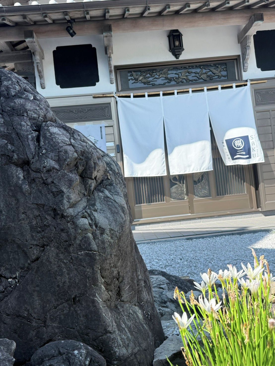 A building with a traditional facade, a large rock in the foreground, and a small garden with white flowers.