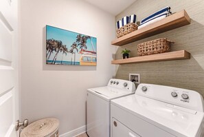 A laundry room with a washing machine, a dryer, and a wall-mounted shelf with baskets. - Low Rates WindMark| Golf Cart + Game Garage Retreat (Port St. Joe)
