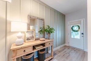 A wooden console table with a mirror, a lamp, and a plant. There are two baskets under the table and a door with a wreath. - SnowBirds Welcome| Low Rates WindMark| Golf Cart + Game Garage Retreat (Port St. Joe)