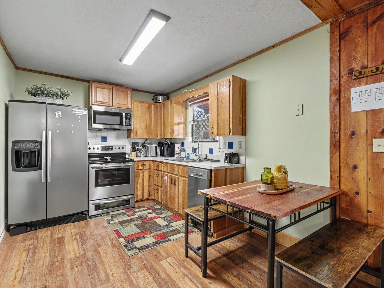 A compact kitchen with wooden cabinets, stainless steel appliances, a wooden table, and a patterned rug.