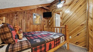 Ein Holzbett mit Etagen, eine gesteppte Bettdecke, ein an der Wand montierter Fernseher und eine Tür auf der rechten Seite.