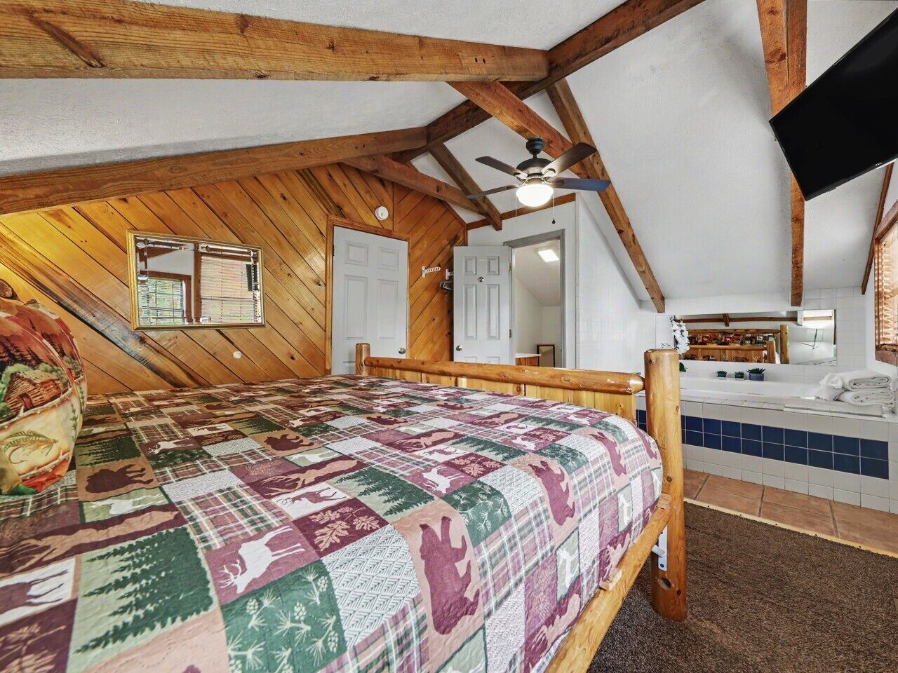 Une chambre à coucher avec un plafond en bois, un lit recouvert d’une courtepointe à motifs, un ventilateur de plafond et une salle de bain visible à travers une porte ouverte.
