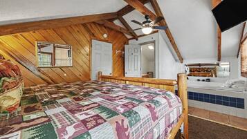 Une chambre à coucher avec un plafond en bois, un lit recouvert d’une courtepointe à motifs, un ventilateur de plafond et une salle de bain visible à travers une porte ouverte.