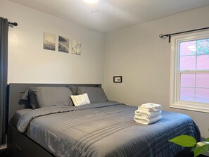 A neatly made bed with a grey duvet and pillows, a window with a view of greenery, and a wall with three framed pictures. - Family-Friendly Getaway for Disneyland Vacation with 2 bedrooms and  2 baths (Santa Ana)