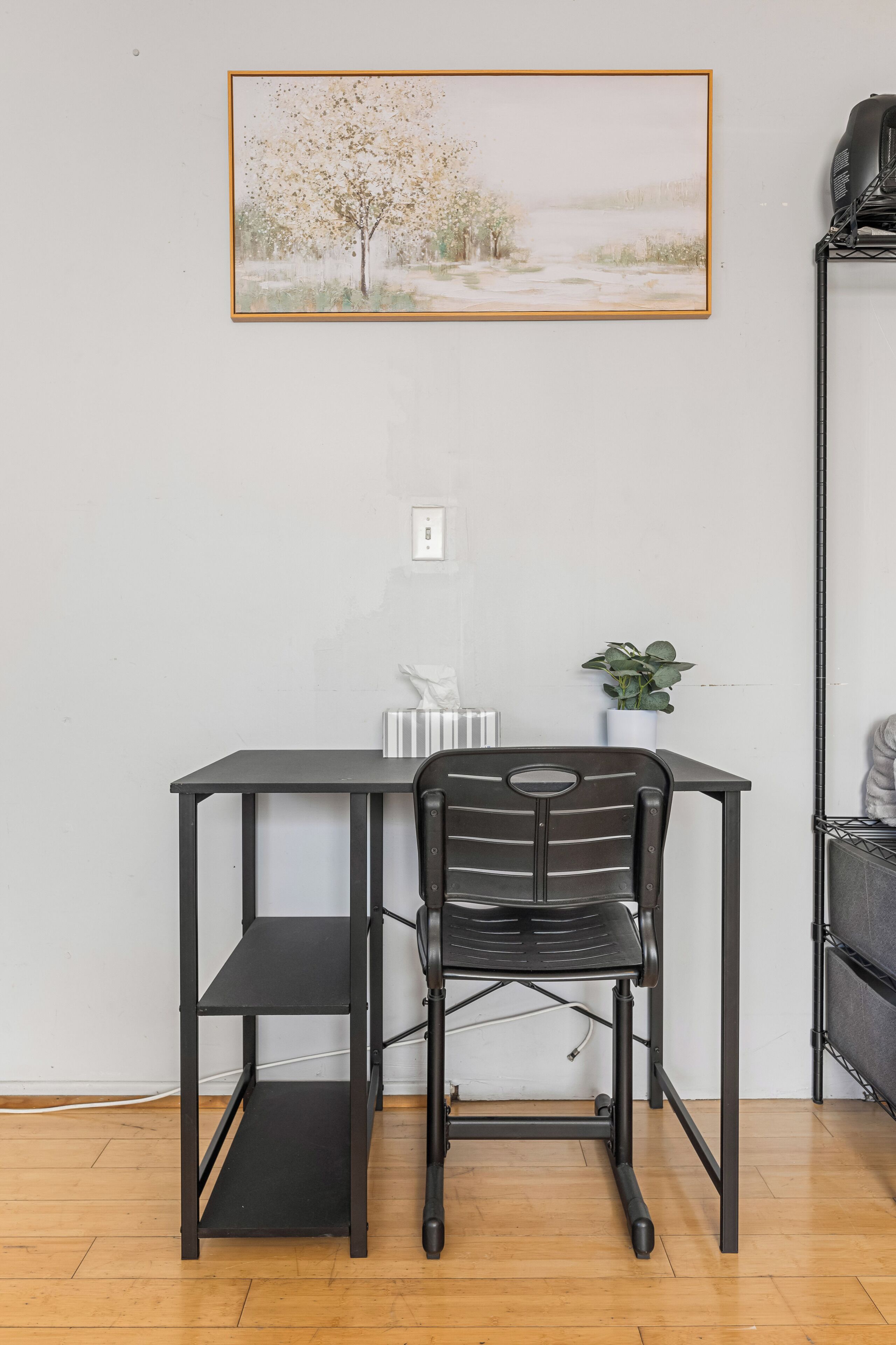 A compact workspace with a desk and chair, a framed painting on the wall, and a potted plant.