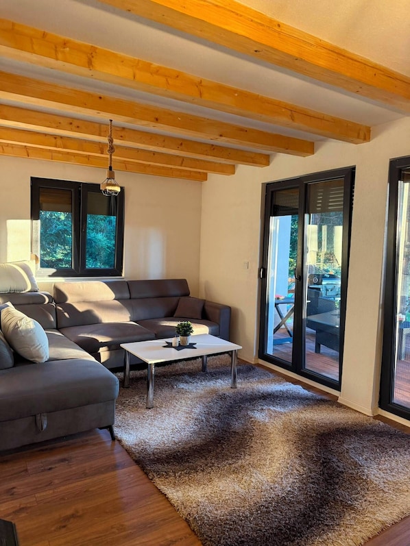 A modern living room with a grey sofa, a white coffee table, and a shaggy rug. There are large windows and a view of the outdoors.