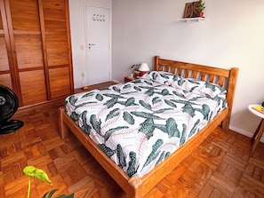 Ein Schlafzimmer mit einem hölzernen Bett, einem Nachttisch, einer Lampe, einem Ventilator, einer Tür und einem kleinen Regal mit einer Pflanze.
