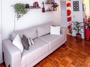 Ein Wohnzimmer mit einer Couch, einer Gitarre, Regalen mit Büchern und Pflanzen sowie einer Wand mit Muster.