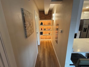 Un pasillo estrecho con suelo de madera, pared de ladrillo y una estantería empotrada con varios objetos.
