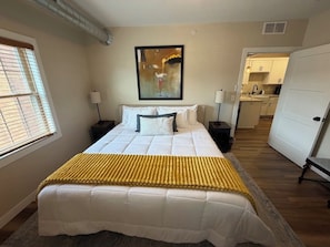 A bedroom with a large bed, a painting on the wall, and a view into the kitchen.