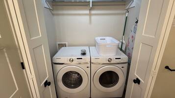 Une buanderie équipée de deux machines à laver Samsung, d’un panier à linge et d’une étagère contenant des produits de nettoyage.