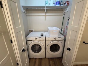 A laundry room with two Samsung washing machines, a basket, and a shelf with cleaning supplies.