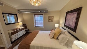 A bedroom with a bed, bedside lamps, a dresser, a window with blinds, and a ceiling vent. - Monroe Lofts Suite 7 Downtown 2BR Apartment (Green Bay)