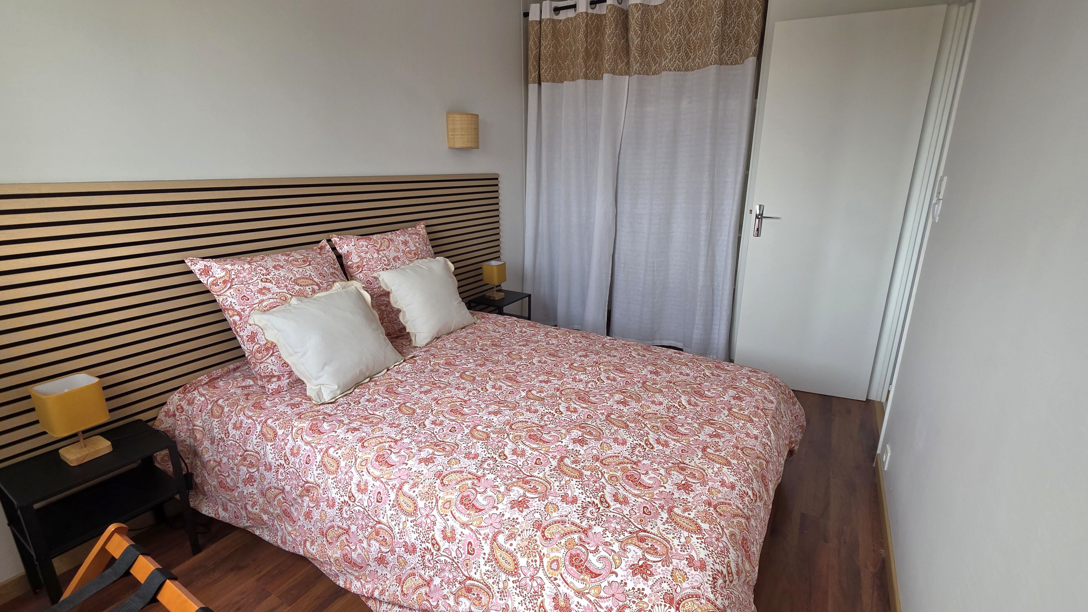 Un lit bien fait, recouvert d’une couvre-lit à motifs, une table de chevet avec une lampe et une fenêtre avec des rideaux.