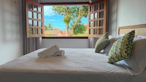 A bed with a view of trees and a building through an open window.
