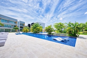 A modern hotel pool area with a clear blue sky, palm trees, and a multi-story building in the background. - Lush Oasis in the heart of 7mile Beach Area (George Town)