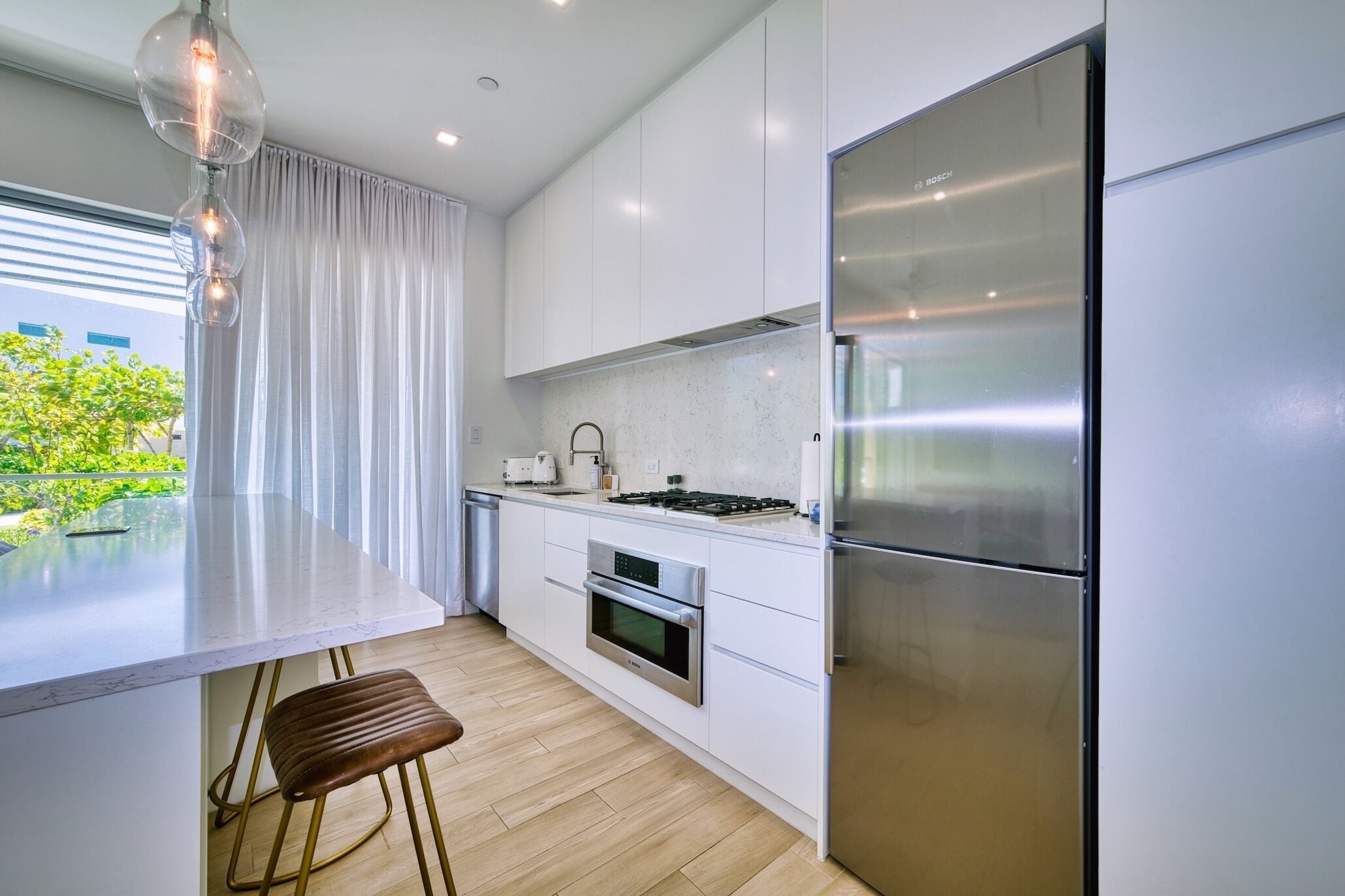 A modern kitchen with white cabinetry, a stainless steel refrigerator, a built-in oven, and a dining area with a bar stool.