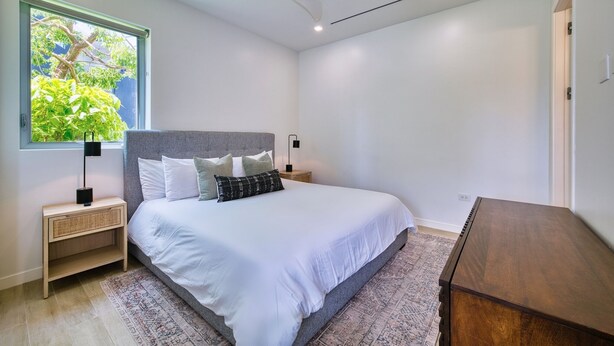 Ein Schlafzimmer mit Bett, Nachttischen, Deckenventilator, Fenster mit Blick auf Grünflächen und einer Holzkommode.