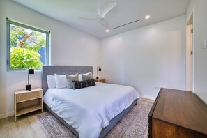 Ein Schlafzimmer mit Bett, Nachttischen, Deckenventilator, Fenster mit Blick auf Grünflächen und einer Holzkommode.
