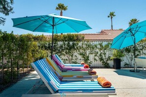 A poolside area with lounge chairs and umbrellas, surrounded by a white fence and palm trees. - Casa Calliandra | Pool, Spa & Game Room (Palm Springs)