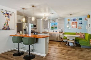 A modern kitchen with a central island, bar stools, and a dining area with a green sofa. - Casa Calliandra | Pool, Spa & Game Room (Palm Springs)