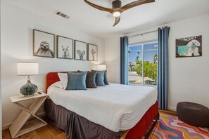 Ein Schlafzimmer mit einem Bett, einem Nachttisch, einer Lampe, einem Deckenventilator und einem Fenster mit Vorhängen.