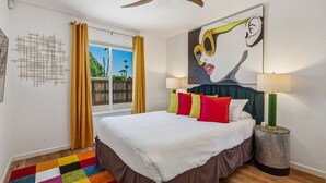 Hotellihuone, jossa on sänky, värikkäitä tyynyjä, taulu, lamppu ja näkymä ulos.