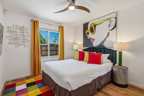 A hotel room with a bed, colorful pillows, a painting, a lamp, and a view of the outdoors. - Casa Calliandra | Pool, Spa & Game Room (Palm Springs)