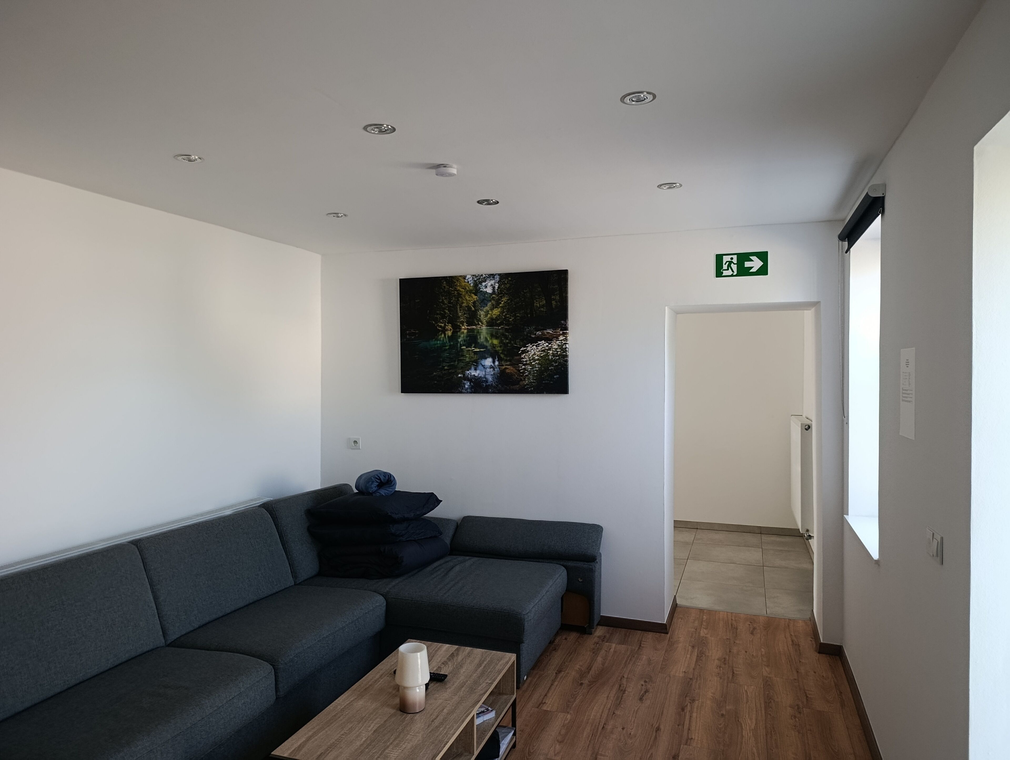 Ein moderner Wartebereich mit einem grauen Ecksofa, einem Holztisch und einem gerahmten Naturfoto an der Wand.