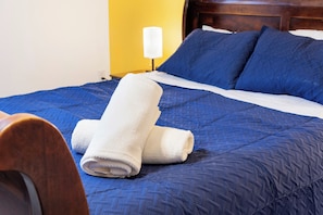 Ein ordentlich bezogenes Bett mit einer blauen Steppdecke, zwei Kissen und einem gefalteten weißen Handtuch.
