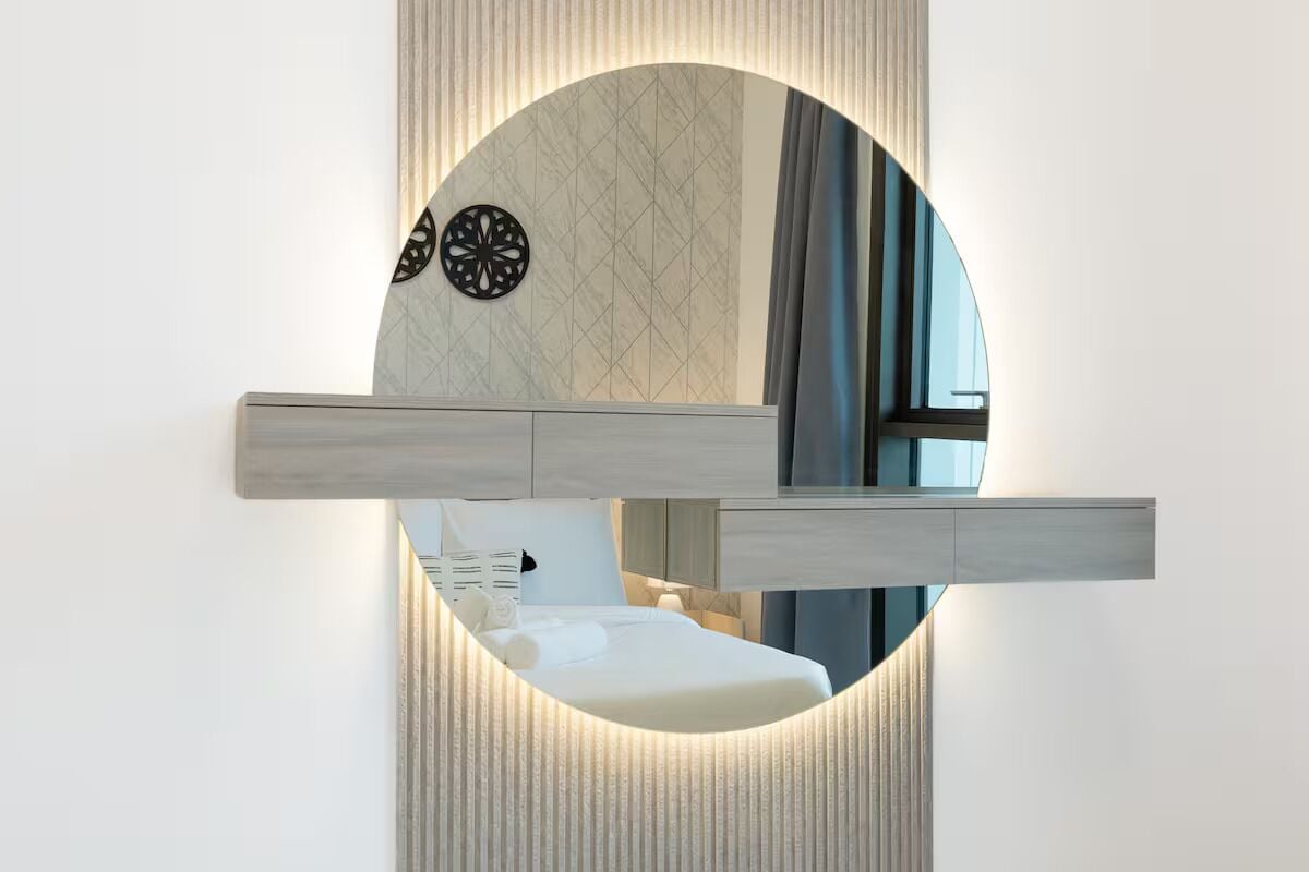 Une salle de bain moderne avec un miroir rond, une étagère en bois et un mur à motifs.