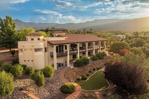 A large, multi-story building with a patio area, surrounded by landscaped gardens and a mountainous backdrop. - Stunning Tuscany Style Home, Breathtaking 360 Views + Casita! (Cottonwood)
