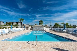 A swimming pool with lounge chairs and umbrellas, surrounded by palm trees and buildings. - Sand Caper #702 (Fort Myers Beach)