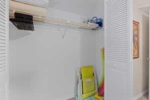 A closet with a white shelving unit, a folded chair, and a door with a picture frame. - Sand Caper #702 (Fort Myers Beach)