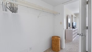A hallway with a white wall, a hanging rack with utensils, a wicker basket, and a door leading to a bathroom with a mirror and a TV. - Sand Caper #702 (Fort Myers Beach)
