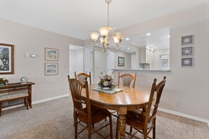 A dining room with a round wooden table, six chairs, a chandelier, and a large mirror on the wall. - Sand Caper #702 (Fort Myers Beach)