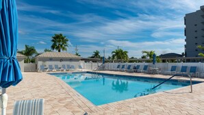 A swimming pool with lounge chairs and umbrellas, surrounded by palm trees and a clear sky. - Sand Caper #702 (Fort Myers Beach)