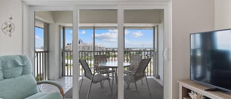 A room with a sliding glass door leading to a balcony with a table and chairs, a television on a wooden stand, and a decorative wall piece above the door.